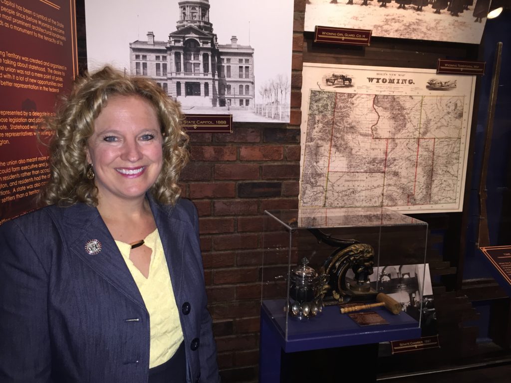 Superintendent Balow smiles while standing near the new display about the Wyoming State Capitol at the State Museum featuring old photographs, maps, a gavel, state seal stamper and other artifacts with descriptions.