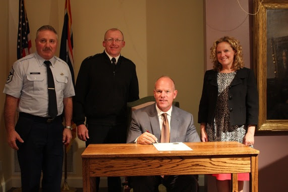 Governor Mead sits at a desk after signing the proclamation as Superintendent Balow, Chief Whipple and General Reiner stand behind him.