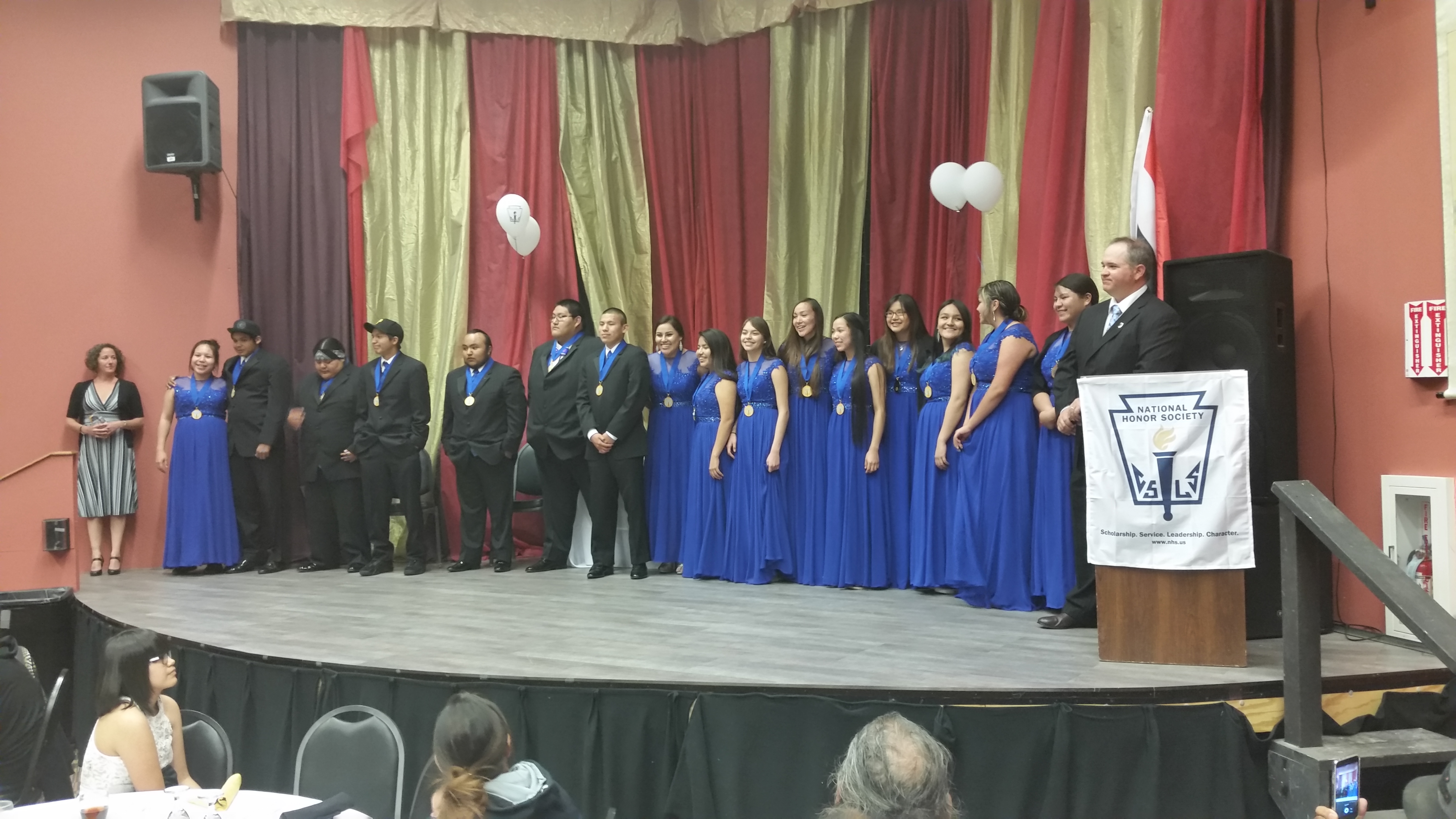 Sixteen students stand smiling on a stage in suits and gowns with their National Honor Society medals around their necks during their induction ceremony.