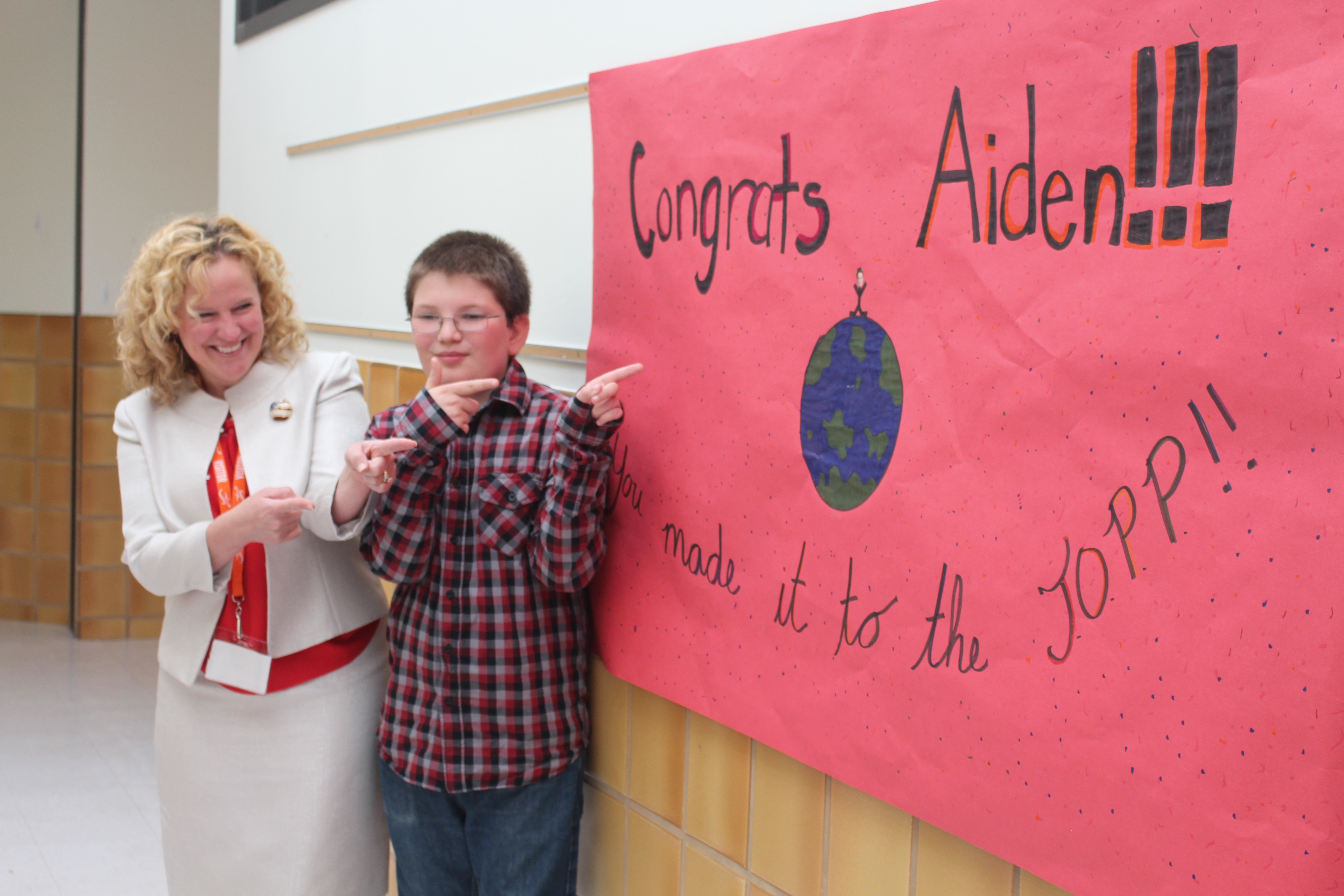 State Superintendent Jillian Balow and Aiden Weinzierl smile and point at a poster that says, "Congrats Aiden!!! You made it to the TOPP!!" and has a stick figure of Aiden sitting on top of the world.