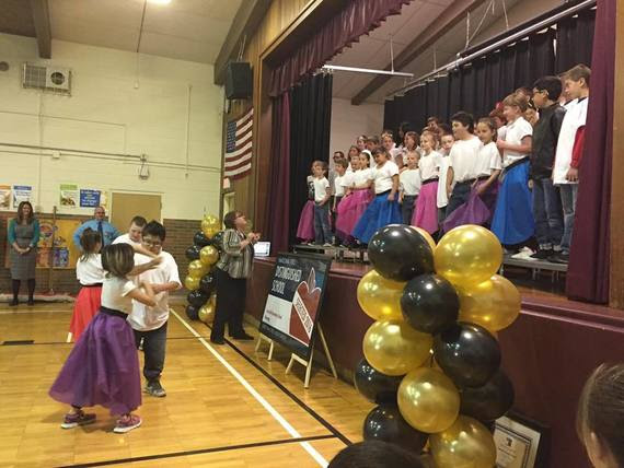 Fourth grade students don poodle skirts and white t-shirts for a performance. Two couples swing dance while their classmates sing on the stage in the school gym during a schoolwide assembly.