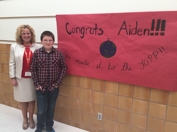 State Superintendent Jillian Balow and winner Aiden Weinzerl stand in front of a poster made by Aiden's classmates which reads, "Congrats Aiden!!! You made it to the TOP!!" and has a stick figure of Aiden standing on top of the world.