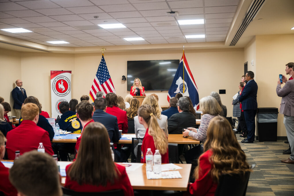 Superintendent Megan Degenfelder stands at a podium while speaking to students who sit at tables facing her.